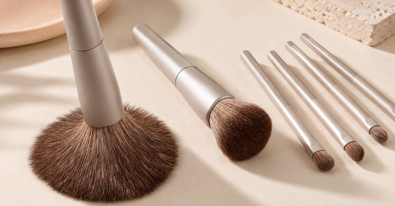 Flat lay of a makeup brush set with various face and eye brushes arranged neatly beside neutral-toned makeup products on a bright vanity surface