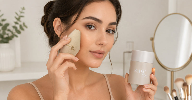 Woman pressing a beige precision blender sponge to her cheek at a vanity with a sponge case beside her